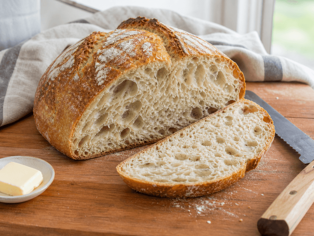 Ein aufgeschnittenes Sauerteigbrot ohne Kneten auf einem Holzbrett mit offener, luftiger Krume und unregelmässigen Löchern. Ein Stück Butter und ein Brotmesser daneben. Die Kruste ist goldbraun mit sichtbarer Mehlbestäubung. Natürliches Küchenlicht.